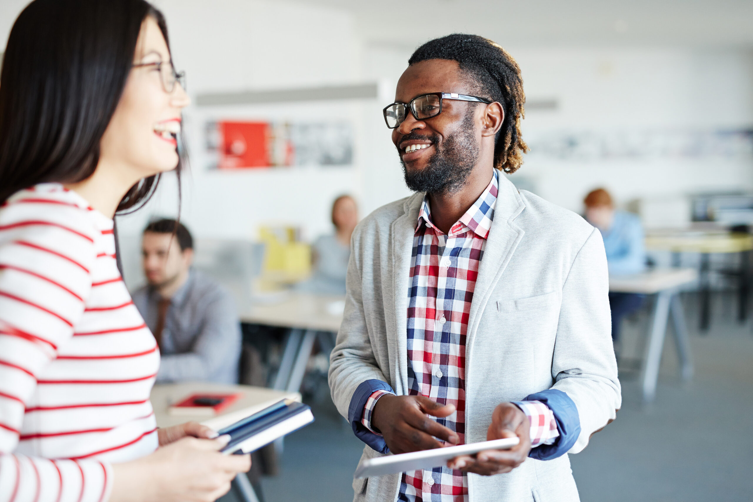 Male and female colleagues standing in open plan office and laughing at something, Afro-American employee holding digital tablet in hands